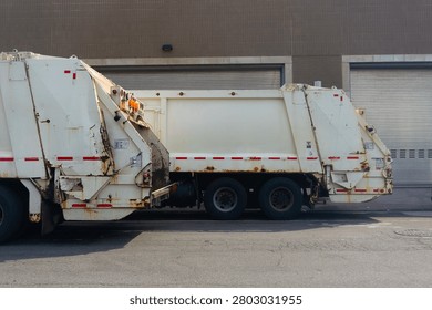 Two rusty garbage trucks are parked on an urban street, showcasing their rear ends with open compartments.
