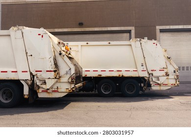 Two rusty garbage trucks are parked on an urban street, showcasing their rear ends with open compartments.
