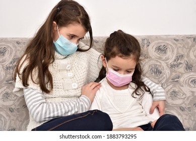 Two sad and depressed sisters sitting on the sofa with medical protective face masks. Older sister hugging her younger sister.