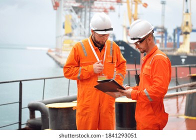 Two sailors in orange overalls work on the ship with documents.