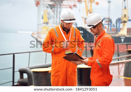 Two sailors in orange overalls work on the ship with documents.