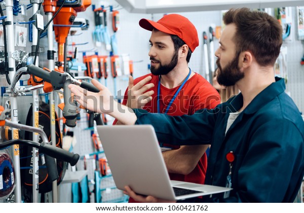 Two\
salesmen in red shirt and baseball cap and blue robe are checking\
tooks inventory with laptop in power tools\
store.