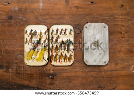 Two salmon fly boxes, one is open showing a selection of old salmon fishing flies . The background is worn brown wood.