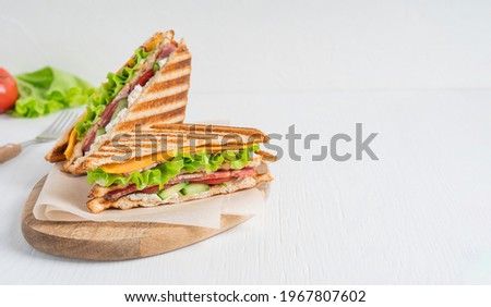 Two sandwiches made of cucumber, slice of meat and cheese, lettuce and tomatoes between slices of grilled bread served on cutting board on white wooden background. Image with copy space, horizontal