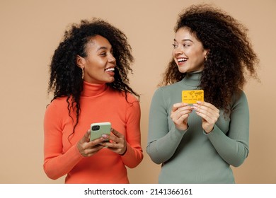 Two Satisfied Young Curly Black Women Friends 20s Wear Casual Shirts Clothes Using Mobile Cell Phone Hold Credit Bank Card Look At Each Other Isolated On Plain Pastel Beige Background Studio Portrait