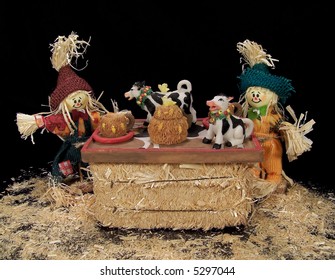 Two scarecrows sitting at a table made of hay having tea from a miniature cow tea set.  Black background.