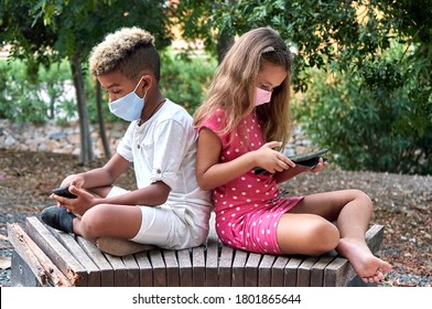 Two school age 9s 10s multi racial kids wear face mask sit together on bench back to back use cellphone and tablet devices ignore each other. Phubbing, new generation addicted with modern tech concept - Powered by Shutterstock