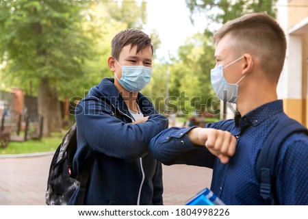 Two school friends greet each other in the schoolyard. Boys maintain a social distance. Their faces are covered in medical masks. They hit each other with their elbows. Concept 2020,
