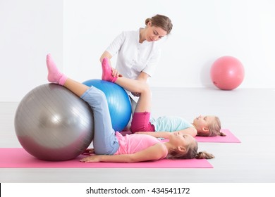 Two School Girls Lying On Exercise Mats Exercising Their Legs On Balls, Accompanied By A Physical Therapist