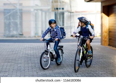 Two School Kid Boys In Safety Helmet Riding With Bike In The City With Backpacks. Happy Children In Colorful Clothes Biking On Bicycles On Way To School. Safe Way For Kids Outdoors To School