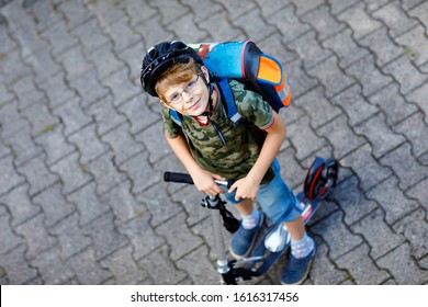 Two School Kid Boys In Safety Helmet Riding With Scooter In The City With Backpack On Sunny Day. Happy Children In Colorful Clothes Biking On Way To School. Unrecognizable Faces From Above Back