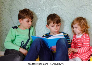 Two School Kids Boys And Little Toddler Girl Reading Together Book. Older Brother Holding Book And Read Aloud For His Sibling. Family Spending Time Together. Three Children.