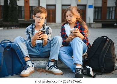 Two school little students small boy and girl sitting on stairs near modern school building. Pretty friends searching internet on smartphone and showing to friend - Powered by Shutterstock