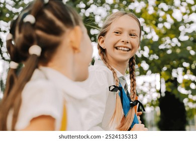 Two schoolgirls with backpacks walk, laugh, and chat outdoors, surrounded by trees, exuding joy and innocence of childhood friendship - Powered by Shutterstock