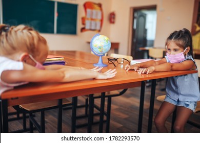 Two Schoolgirls In Medical Masks Are Sitting At A School Desk, Opposite Each Other, Group Session, Back To School, Teaching Children, Social Distance During Epidemic.