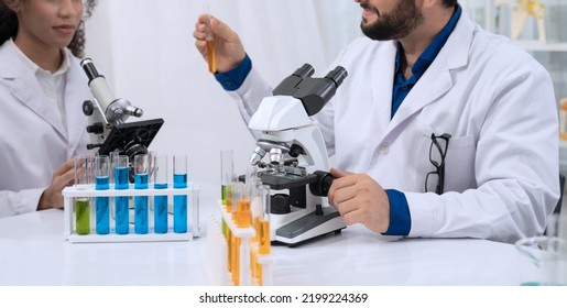 Two Scientist Researchers Wearing White Coat And Working In Laboratory. Man Holding Test Tube With Yellow Liquid And Using Microscope. Chemistry, Medical Biology And Phamacy Reseach Concept.