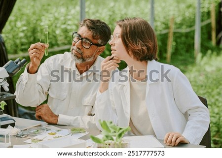 Two scientists analyzing plant samples outdoors, holding test tube and discussing results, surrounded by laboratory equipment and greenery under a shade structure