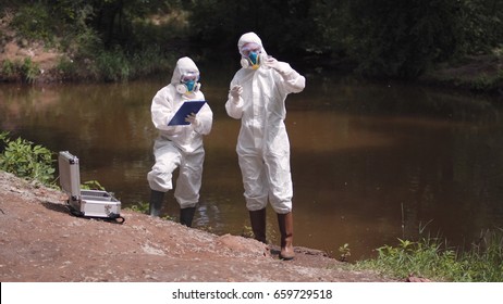 Two scientists in full biohazard suits and masks taking water samples from a river to check for hazardous pollutants or contaminants