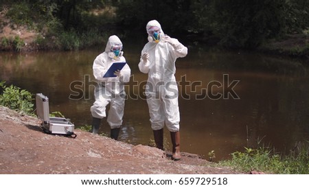 Two scientists in full biohazard suits and masks taking water samples from a river to check for hazardous pollutants or contaminants