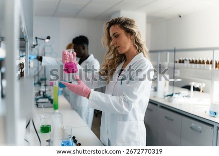 Two scientists in lab coats and gloves examining a pink chemical liquid in a beaker, conducting research in a modern laboratory filled with equipment and solutions