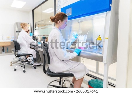 Two scientists in lab coats work in a clean, contemporary laboratory. One focuses on samples under a fume hood, while the other analyzes data at a desk.