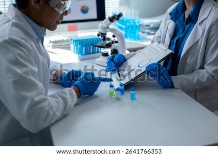 Two scientists are looking at a chart and pointing at it. They are wearing lab coats and gloves