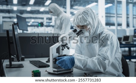Two Scientists In Sterile Suites At Electronics Factory: Engineer Using Microscope And Desktop Computer To Inspect Microchip While His Colleague Using Soldering Jet Printer To Print Modern Motherboard