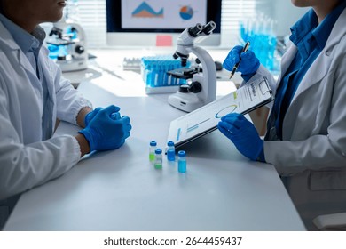 Two scientists wearing lab coats and gloves are examining data and vials in a laboratory