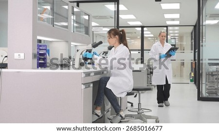 Two scientists work in a well-lit laboratory. One is using a microscope while the other examines data on a tablet. The lab is organized with modern equipment and a clean environment.