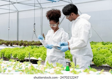 Two scientists are working in a greenhouse, one of them is wearing a white lab coat and the other is wearing a white lab coat and a pair of gloves. They are looking at a plant - Powered by Shutterstock