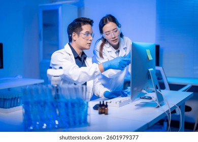 Two scientists are working on a computer. One of them is wearing a lab coat. The computer monitor is on the desk