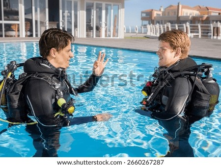 Two scuba divers in a pool at a dive resort in full wetsuits and scuba gear. Instructor showing the ok signal to a smiling student