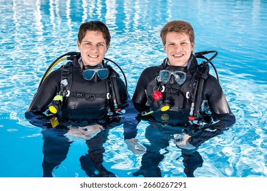 Two scuba divers in a pool, looking at the camera, smiling, wearing full wetsuits and scuba kits with their dive masks around their necks.