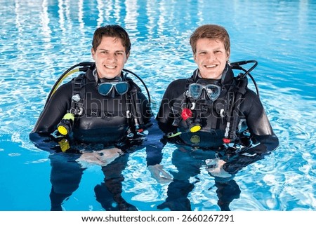 Two scuba divers in a pool, looking at the camera, smiling, wearing full wetsuits and scuba kits with their dive masks around their necks.
