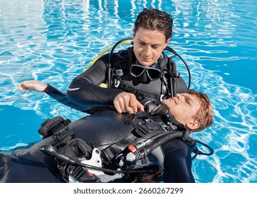Two scuba divers practicing diver rescue skills in a pool, with one diver floating on his back and the other is unclipping his BCD