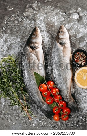 Two seabass, which are located on ice particles on the kitchen table in one direction, next to which there is a black pot with pepper, sprigs of fragrant rosemary, a branch with cherry tomatoes