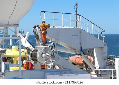 Two Seafarers Painting Ship's Crane While Out At Sea. Both Wearing Full PPE And Fall Protection Harnesses To Prevent Fall And Injury. Onboard Safety Culture Concept.