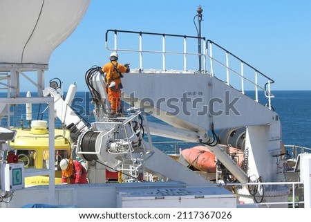 Two seafarers painting ship's crane while out at sea. Both wearing full PPE and fall protection harnesses to prevent fall and injury. Onboard safety culture concept.
