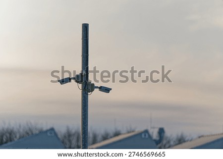 Two security cameras attached to a tall metal pole monitoring an outdoor area with buildings and trees in the distance under a cloudy sky.