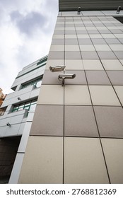 Two security cameras are fixed on the side of a modern building. The cameras watch over the city area under a cloudy sky in daylight.