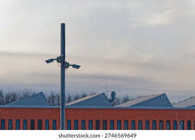 Two security cameras are installed on a tall metal pole monitoring an industrial area with a brick building and metal rooftops under a cloudy sky at dusk.
