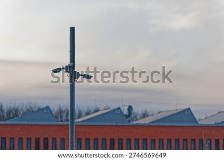 Two security cameras are installed on a tall metal pole monitoring an industrial area with a brick building and metal rooftops under a cloudy sky at dusk.