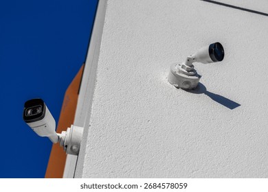 Two security cameras mounted on a building corner monitor the area, capturing footage against a bright blue sky.