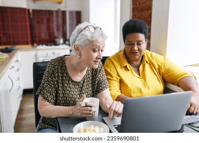 Two senior best female friends or neighbors sitting at kitchen table in front of laptop drinking tea and watching series of show together, discussing and arguing. Friendship for life - Powered by Shutterstock