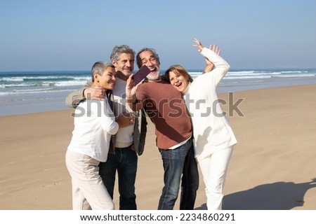 Two senior couples having fun at the beach, standing against sea background and taking selfie. Excited bearded man holding smartphone and making shot. Modern technology, friendship, leisure concept