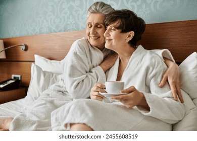 Two senior lesbians in robes share a tender moment on a bed, one holding a cup. - Powered by Shutterstock