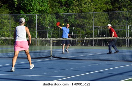 Two Senior Men Double Up Against A Woman In A Competitive Game Of Pickle Ball.  They Are Playing On A Tennis Court.