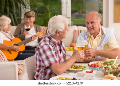 Two senior men drinking beer and their wives playing guitar during dinner