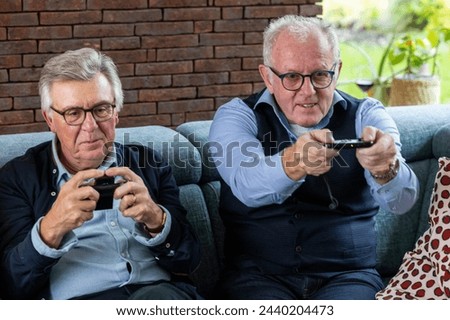 Two senior men exhibit concentration and enjoyment while playing video games, seated comfortably on a couch. A brick wall provides a warm backdrop, highlighting the modern pastime within a homely