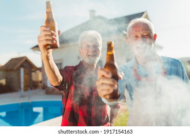 Two Senior Men Making A Toast With Bottles Of Beer While Grilling Meat At Backyard Barbecue Party By The Swimming Pool, Relaxing Outdoors On Sunny Summmer Day